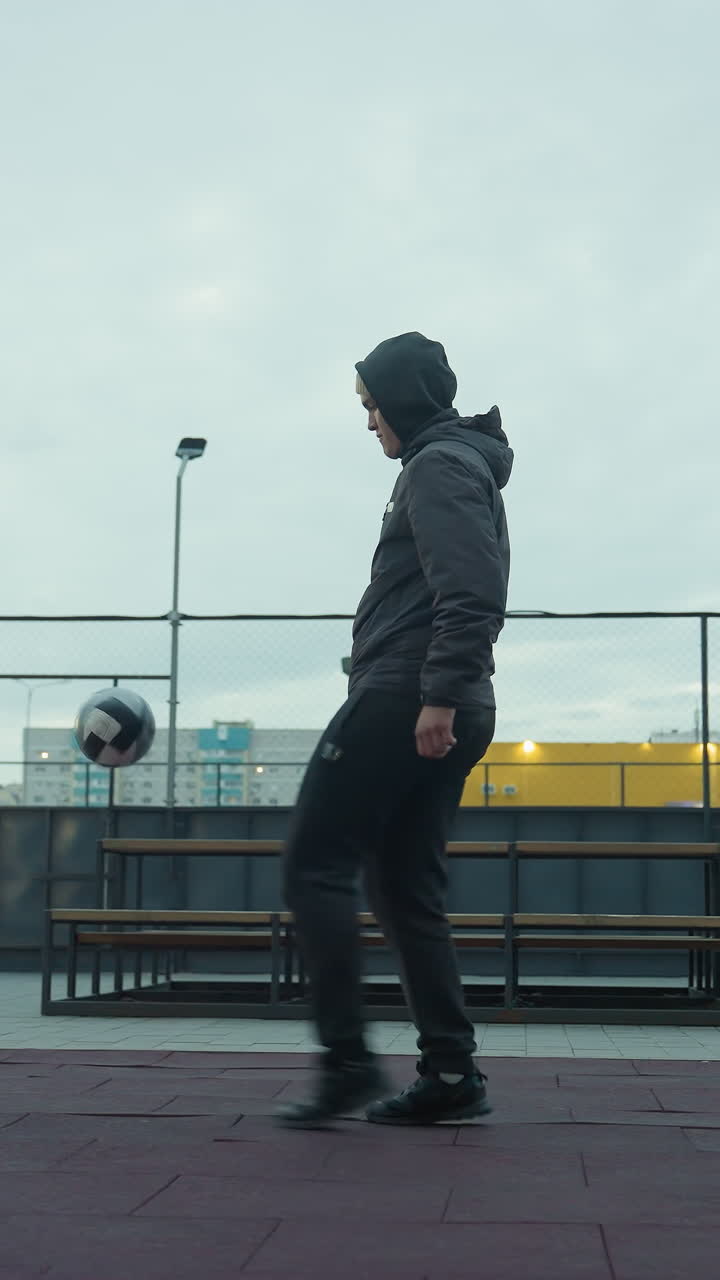 Man skillfully juggling football on sport arena with urban background featuring benches, lit pathways, and tall residential buildings