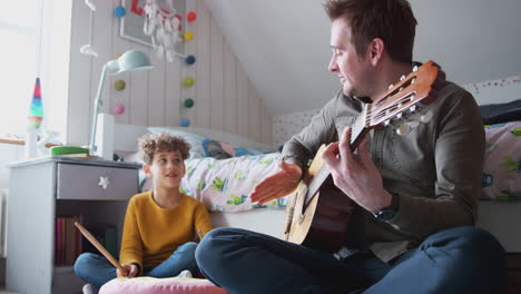 single father playing guitar with son who drums on cushion in bedroom