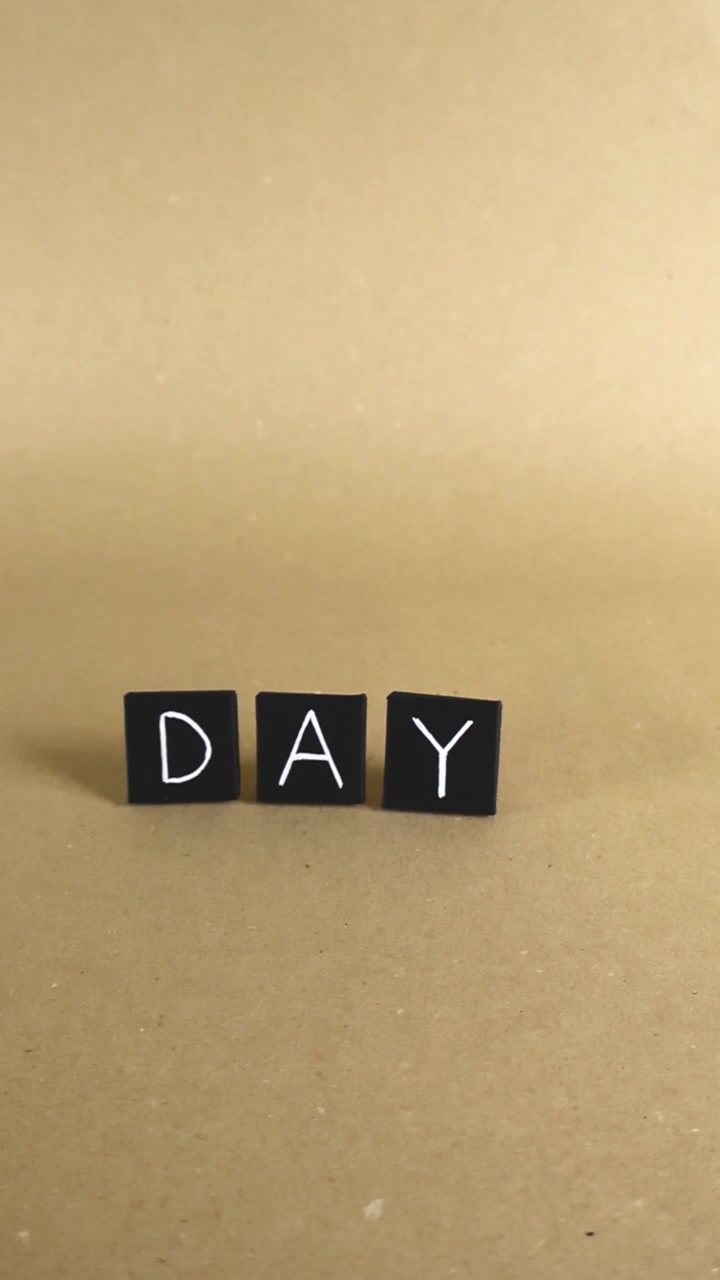 Piastrelle di carta nere con lettere bianche che formano la parola "day" scorrono su uno sfondo di cartone marrone chiaro, a simboleggiare il passare del tempo, le routine quotidiane e i nuovi inizi.