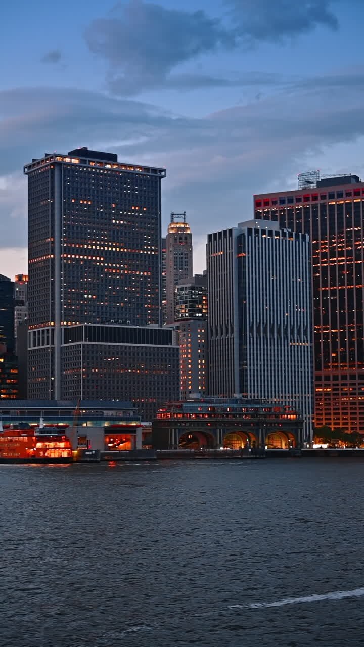 Diverse high-rise buildings with lights on at the waterfront of the East River. Modern skyline of New York, USA at dusk time from the river side. Vertical video