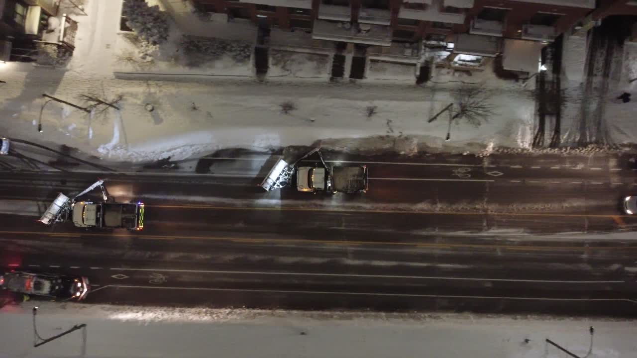 Snow plows work to clear slushy snow from an urban street at night, surrounded by buildings, parked cars, and tire tracks on wet asphalt, viewed directly from above during active snowfall.