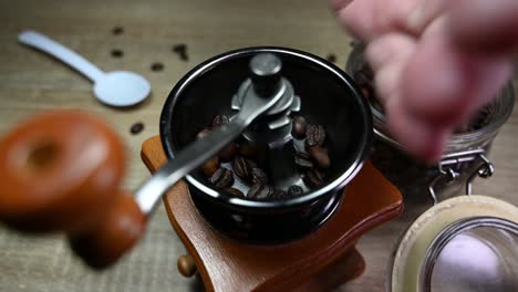 Mano-Izquierda-Tomando-Granos-De-Café-Del-Frasco-Y-Llenando-El-Molinillo-Mientras-Algunos-Granos-Caen-Sobre-La-Mesa