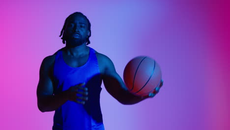 studio portrait of male basketball player wearing team vest bouncing ball against colourful low key mixed lighting 1