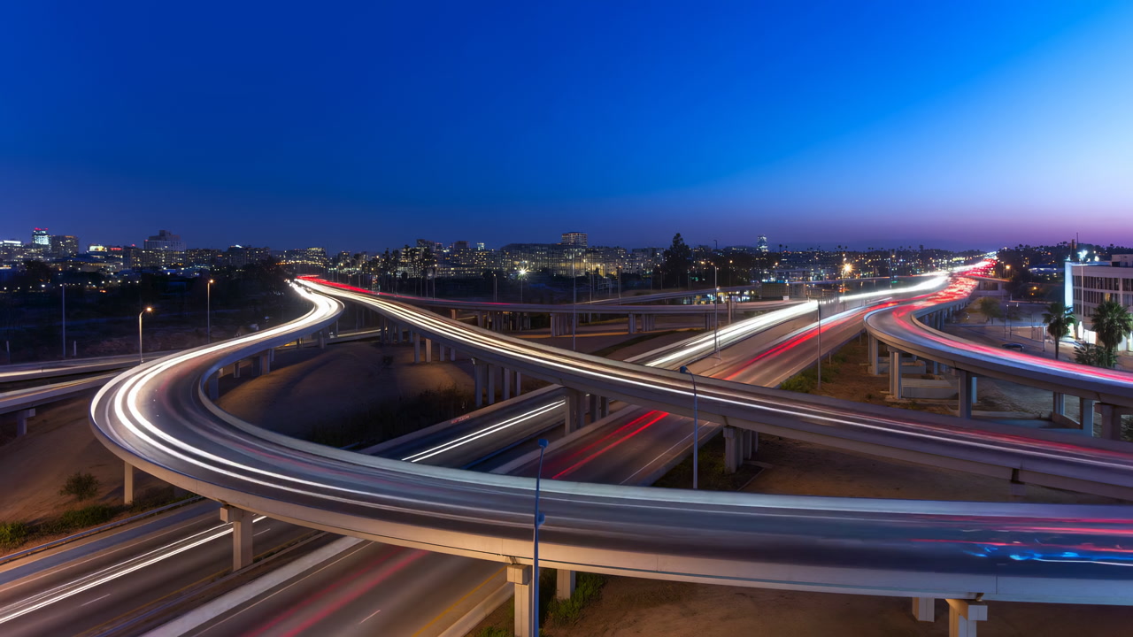 Night Highway Interchange With Sweeping Light Trails Across Curved Overpasses And Distant City Skyline, Long