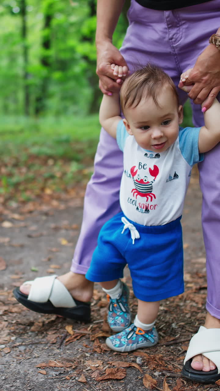 Mom's holding her baby's hands helping him to walk. Little child learning to walk outdoors. Nature backdrop. Vertical video