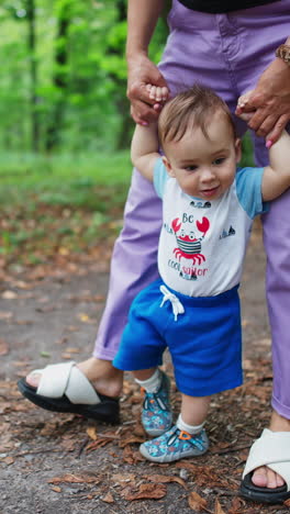 mom's holding her baby's hands helping him to walk. little child learning to walk outdoors. nature backdrop. vertical video