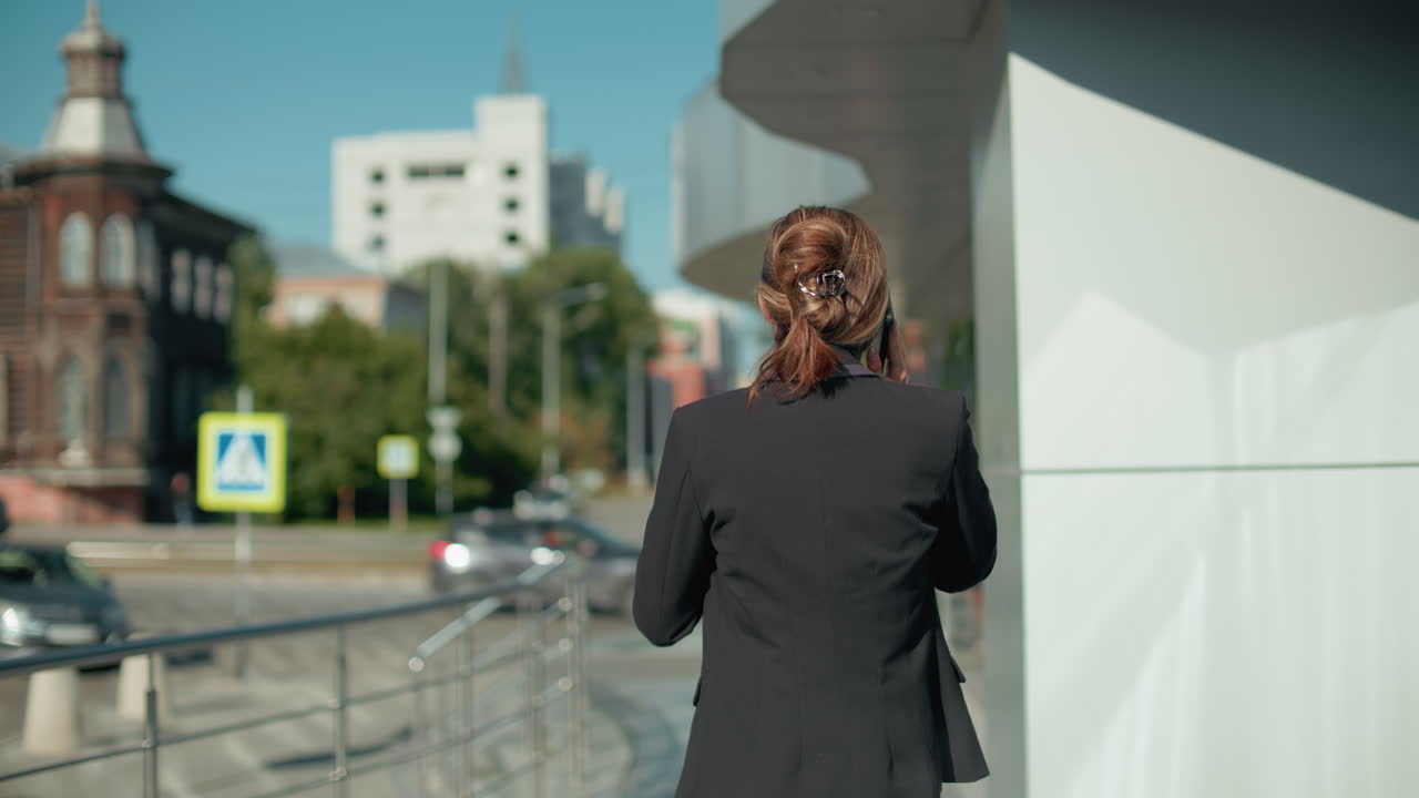 Back view of woman in black suit with hair tied back on phone call sipping coffee while walking along urban sidewalk next to building and iron railing with blurred background traffic and signage