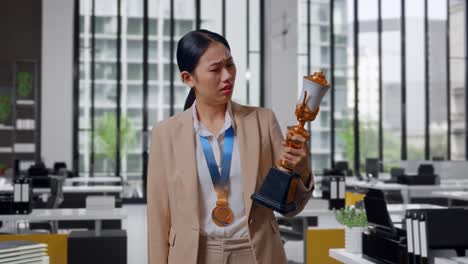 mujer de negocios asiática molesta en un traje con una medalla de oro mirando el trofeo luego sacudiendo la cabeza siendo insatisfecha ganando como la primera ganadora en la oficina