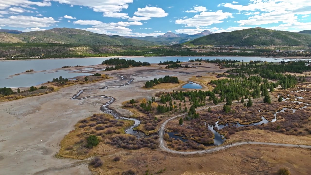 Ampia veduta aerea di una regione montuosa con lago e fitte aree boschive sotto un cielo blu, un fiume serpeggiante e prosciugato si snoda attraverso il paesaggio