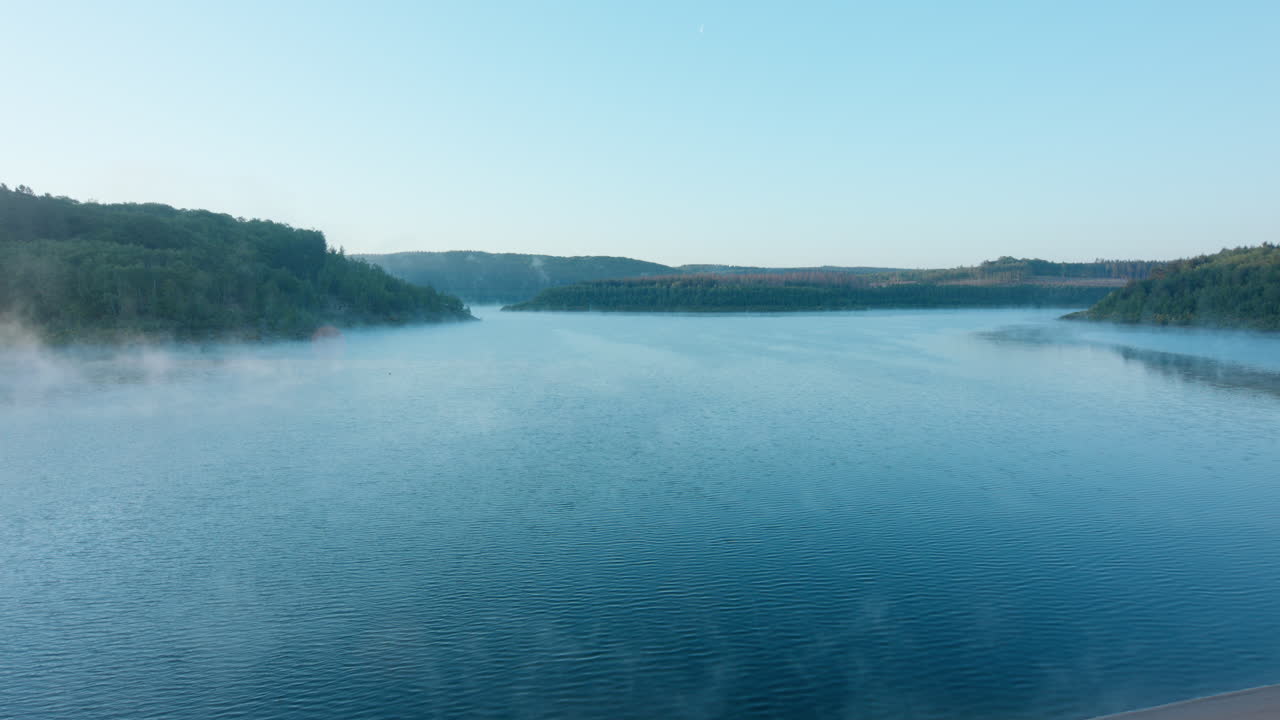 High quality video showing a calm lake at dawn with soft mist drifting across the water and gentle morning light over distant hills