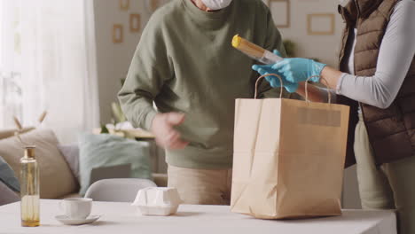 woman in mask and gloves unloading groceries for senior man at home