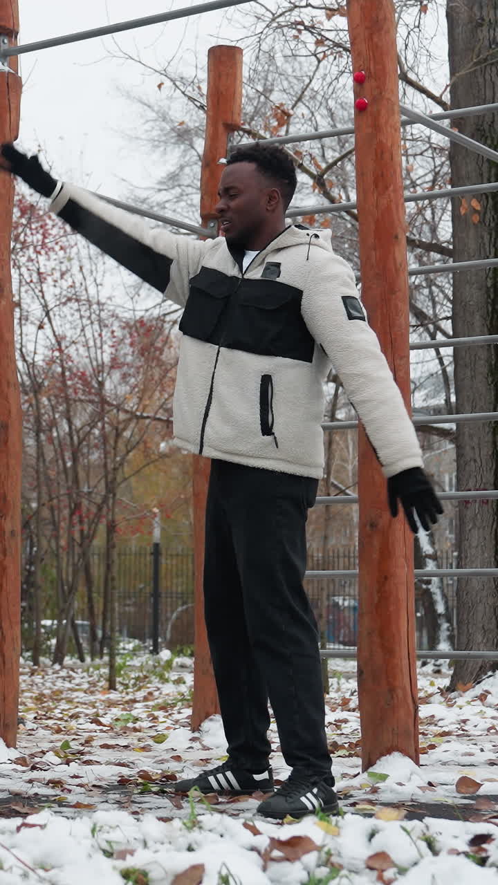joven extiende los brazos para la flexibilidad en el área de entrenamiento de invierno al aire libre, vestido con lana cálida y guantes, hace ejercicio cerca de equipos de fitness de madera con árboles, suelo cubierto de nieve