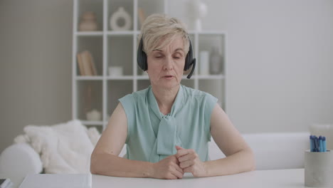 aged woman is speaking to headphones with microphone and looking at camera sitting at table in home call-center and videoconferencing technology