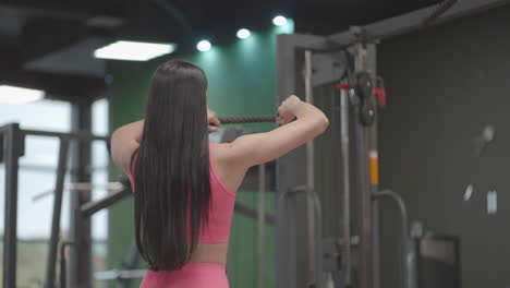 a brunette woman in a pink suit pulls a rope in a crossover trainer to her chest. back workout in a trainer. professional woman instructor