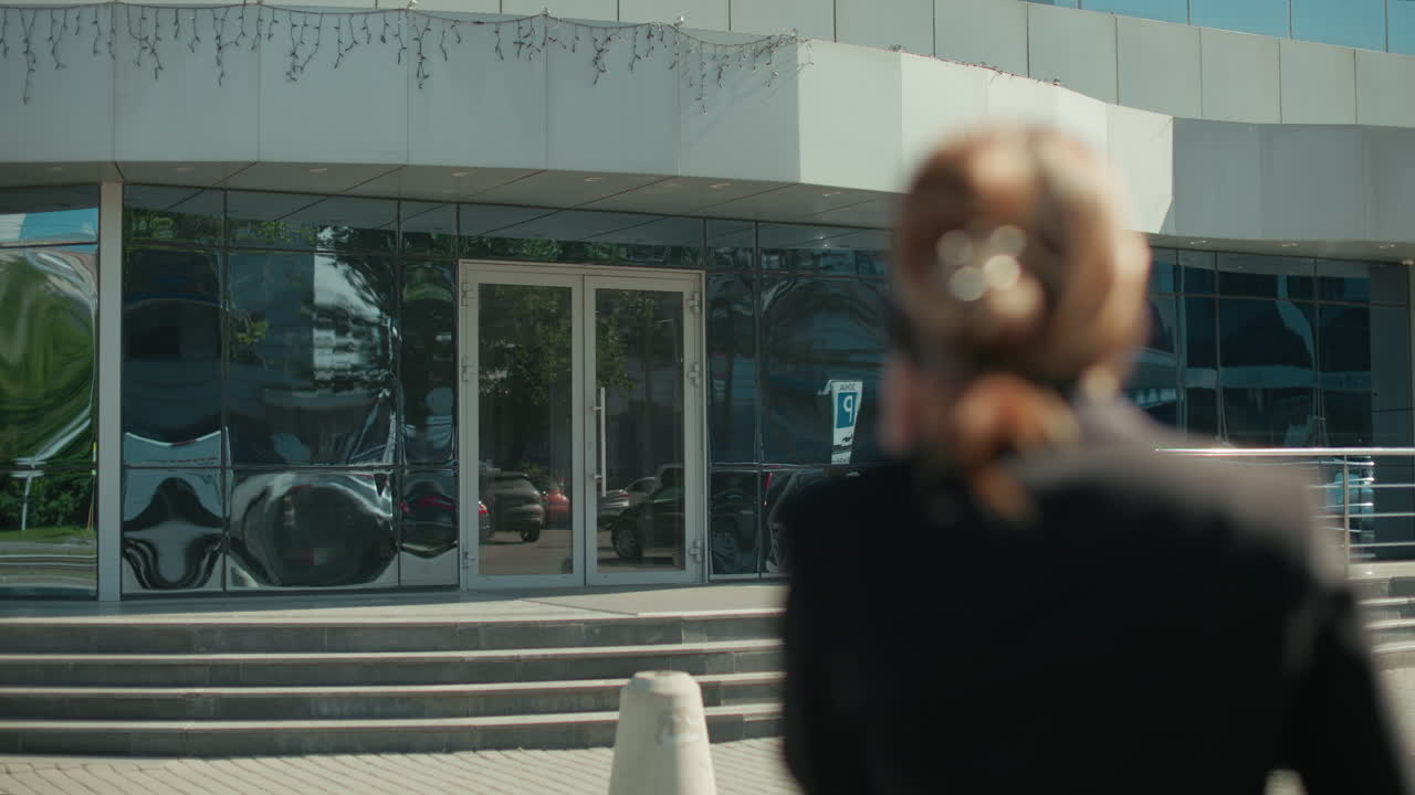 Back view of professional woman holding handbag walking towards work on phone call, with modern building reflecting parked cars in glass facade