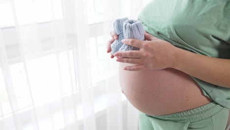 unidentified pregnant woman paces small baby socks on the top of her big tummy and moves them slowly downwards. future mom plays with knitted baby booties. close up.