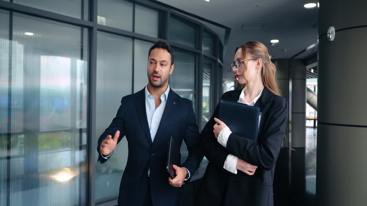 Confident experienced boss explains project details and gives constructive feedback to focused female employee while walking together in modern office corridor