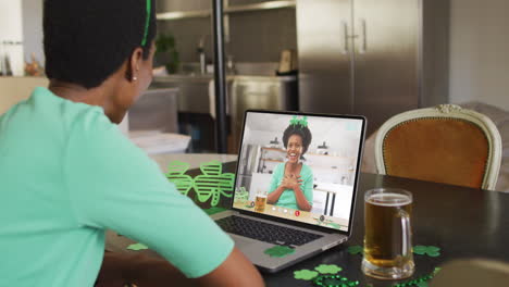 mujer afroamericana sonriente con cerveza vistiendo banda en forma de trébol en una llamada de video en una computadora portátil