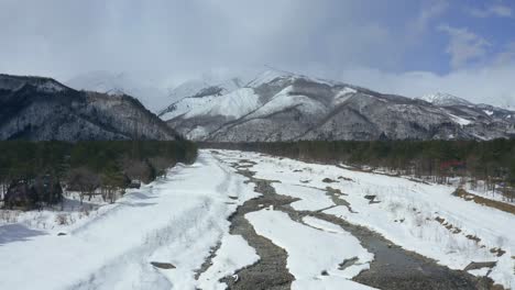 Deslizándose-Hacia-Adelante,-El-Dron-Captura-La-Belleza-De-Un-Río-Invernal-En-El-Valle-De-Hakuba,-Japón,-El-Flujo-Helado-Se-Abre-Paso-A-Través-De-Las-Orillas-Nevadas-En-Un-Hermoso-Paisaje-Montañoso.