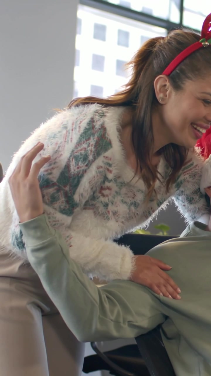 Video verticale: Donna che arriva, con maglione e corna, si china su un collega al lavoro sistemando un cappello da Babbo Natale con allegria