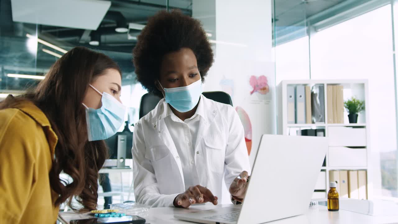 Close-up view of African American female doctor in medical mask using the laptop and explaining to female patient treatment for coronavirus in medical consultation