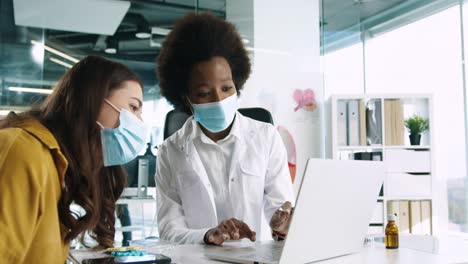 Close-up-view-of-African-American-female-doctor-in-medical-mask-using-the-laptop-and-explaining-to-female-patient-treatment-for-coronavirus-in-medical-consultation