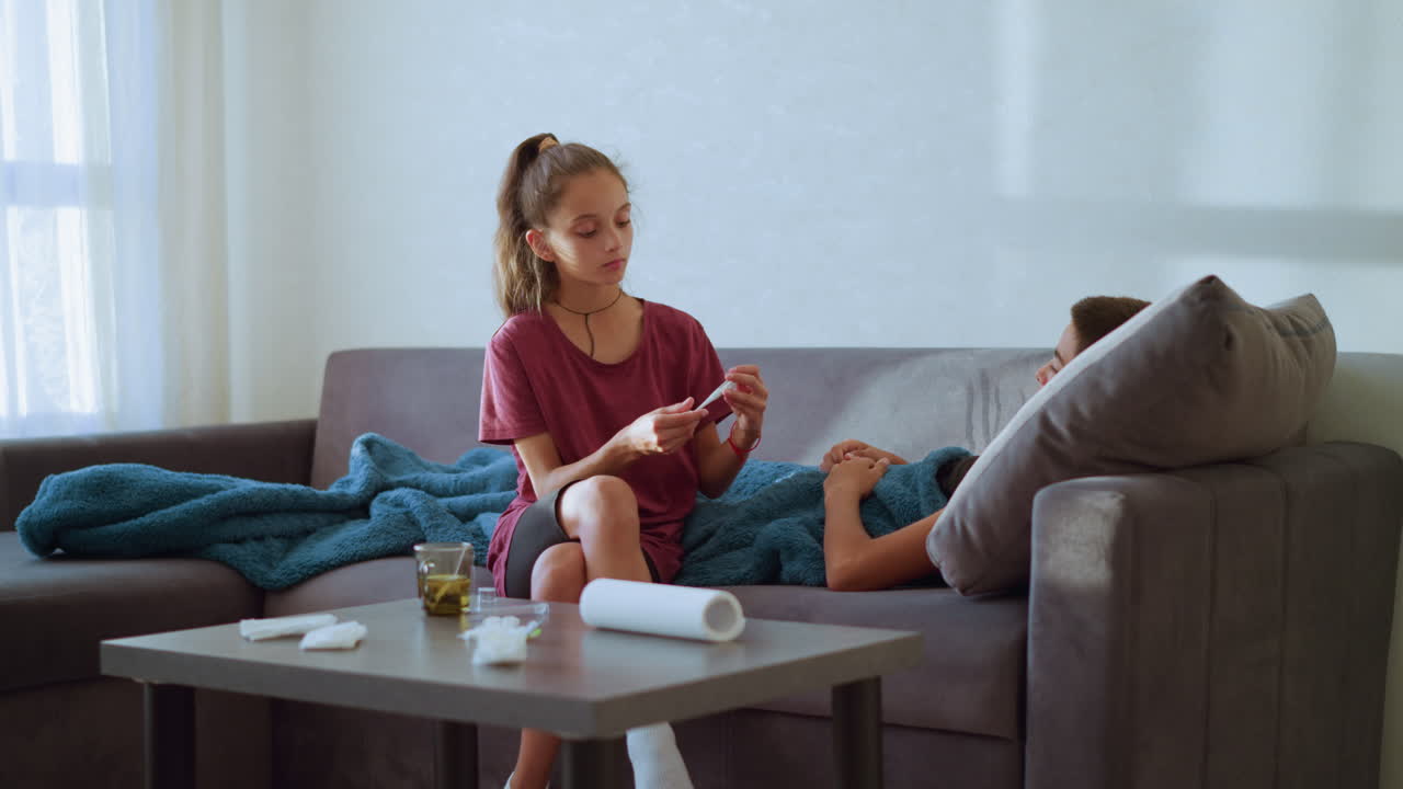 Young girl sits cross-legged on couch checking thermometer used by sick brother while holding his hand showing care and concern with tissue paper and glass cup on table