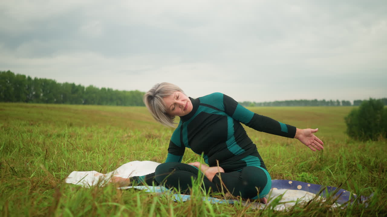 회색 머리카락의 여자가 요가 매트에 앉아서 멀리 나무로 둘러싸인 푸른 하늘 아래 광활한 잔디에서 손을 펴고 이 구부리는 자세를 연습하고 있습니다.