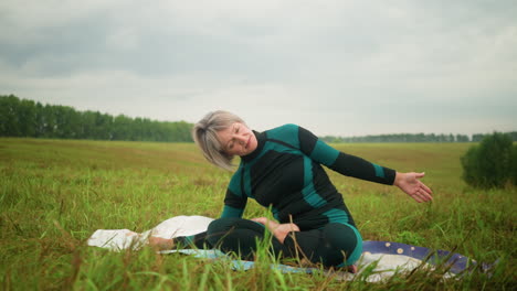 woman with grey hair seated on yoga mat practicing side bend pose with hands stretched in a vast grassy field under cloudy skies, surrounded by nature with trees in the distance