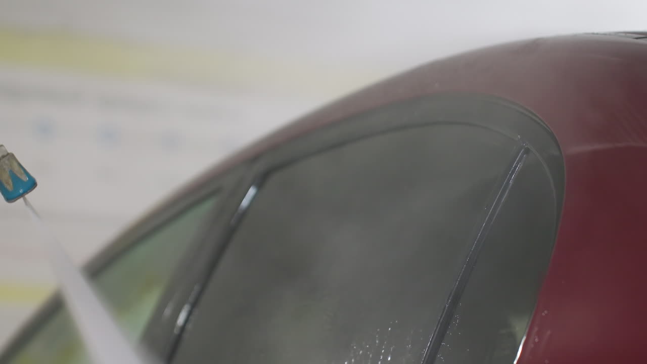 Macro close up of high pressure water rinsing red car side window with suds residues and dynamic droplets streaming over glass surface inside professional indoor garage during car wash service