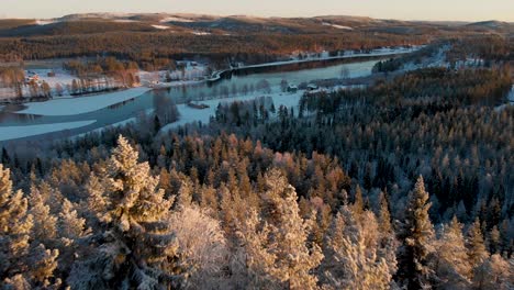 Vista-Aérea-De-Un-Pequeño-Pueblo-En-La-Distancia-Rodeado-De-Pinos-Escandinavos-Cubiertos-De-Nieve-Durante-La-Puesta-De-Sol