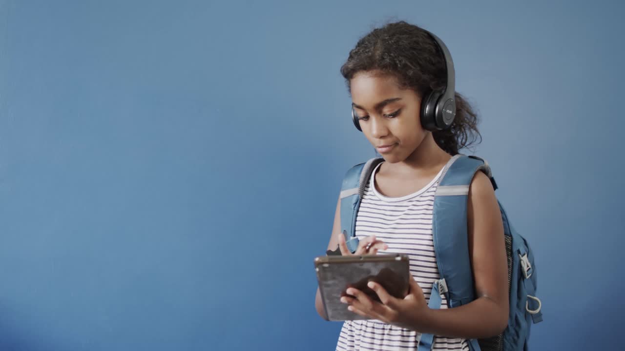 Happy african american girl using headphones and tablet on blue background, copy space, slow motion