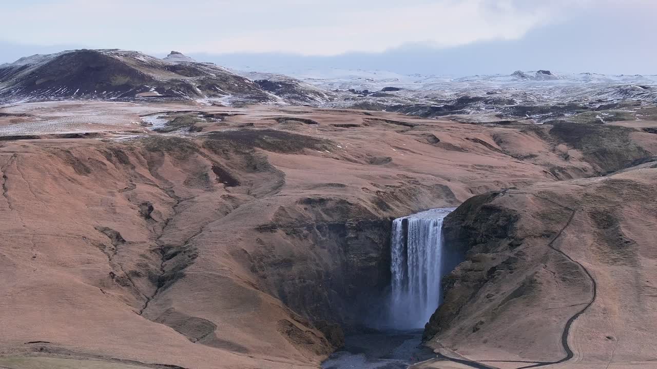 Wide aerial view of Skógafoss waterfall flowing through the rugged Icelandic landscape near Eyjafjallajökull, with snowy mountains in the background.