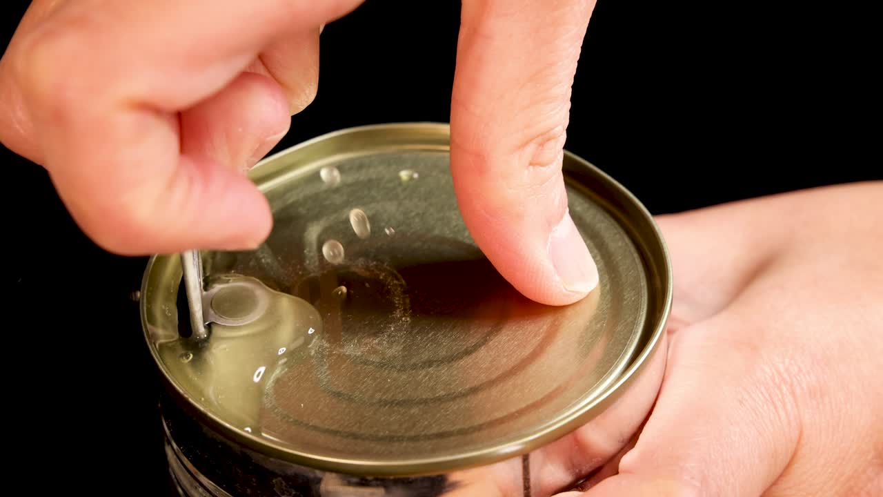 A person’s hands open a canned beans container using a pull tab, shown in close-up with even lighting and a black background