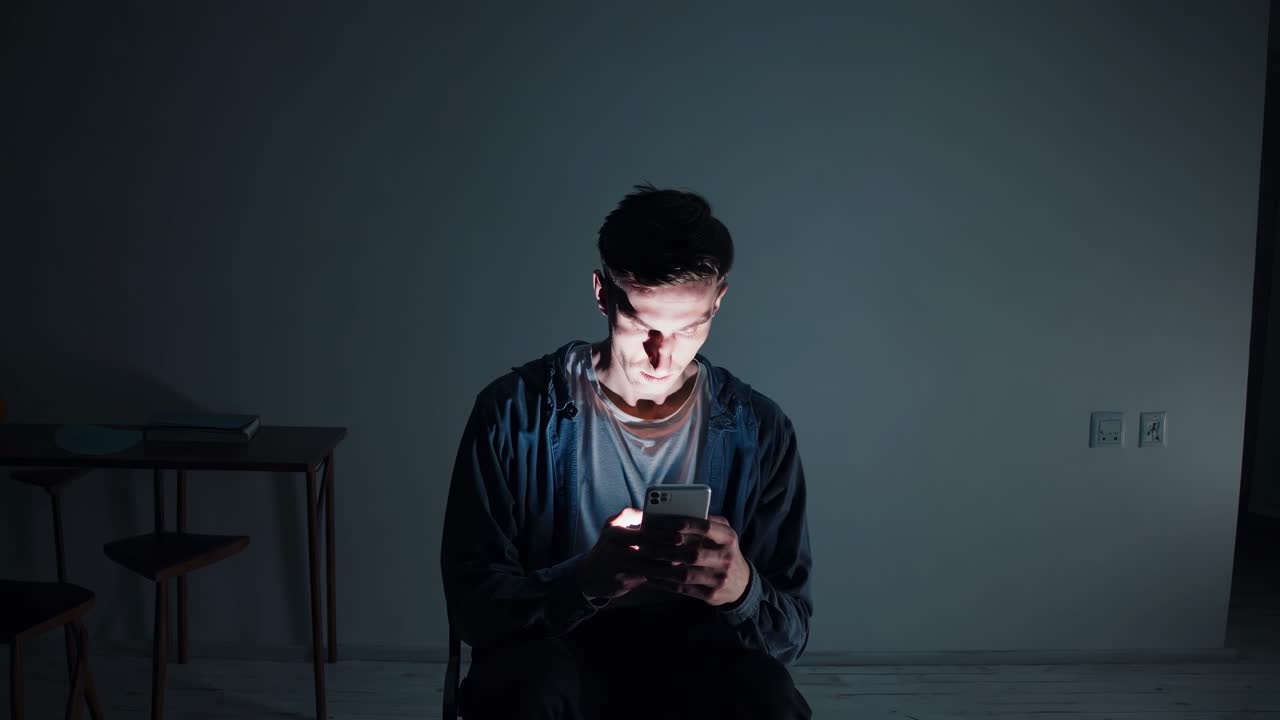 Young man seated in a dimly lit room, focused on his smartphone, showcasing the evolving emotions and engagement with digital technology in a serene atmosphere