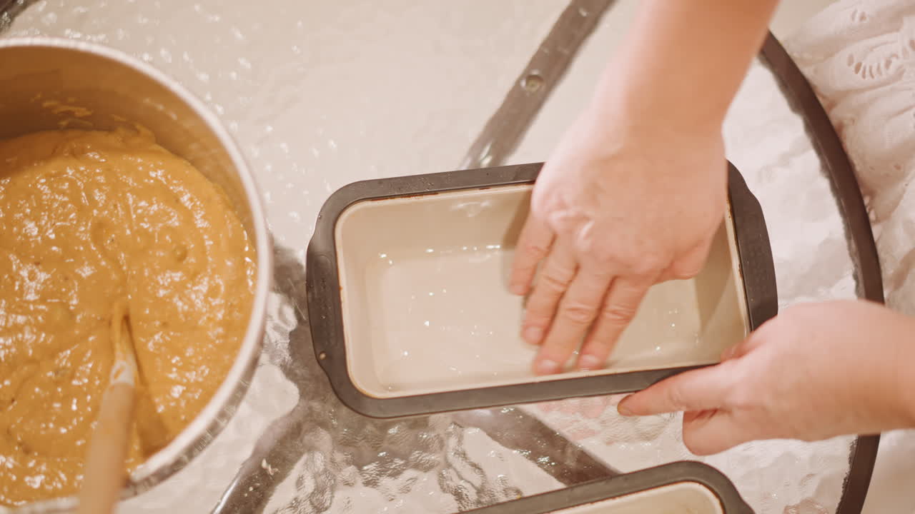 Greasing loaf pan with hand overhead shot of baker pressing butter and flour into ceramic loaf tin, smoothing parchment lining, preparing multiple tins for spiced pumpkin bread, careful prep and neat