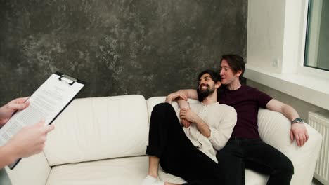 Happy-gay-couple-with-real-estate-agent-relaxing-on-sofa,-excited-about-their-new-apartment