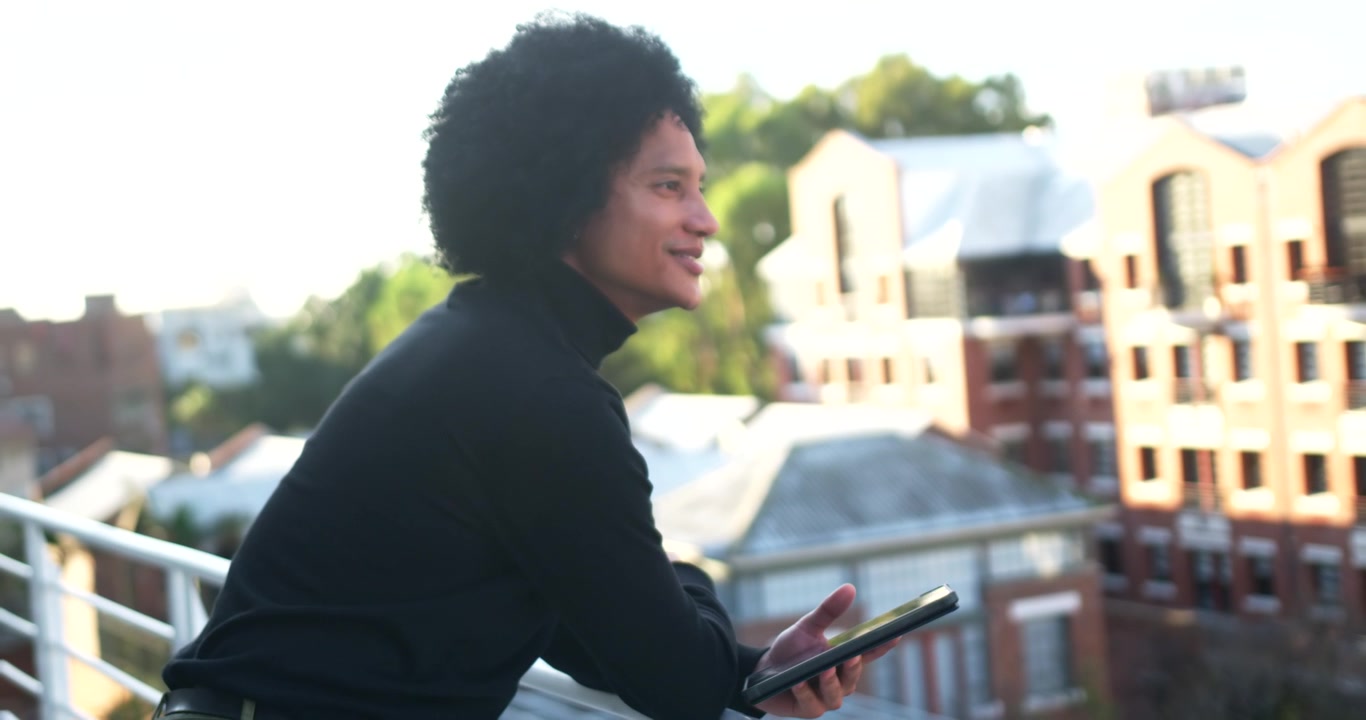 Afro-Amerikaanse man leunt op dakreling met tablet, kijkt naar tablet, tilt kin op, pauzeert