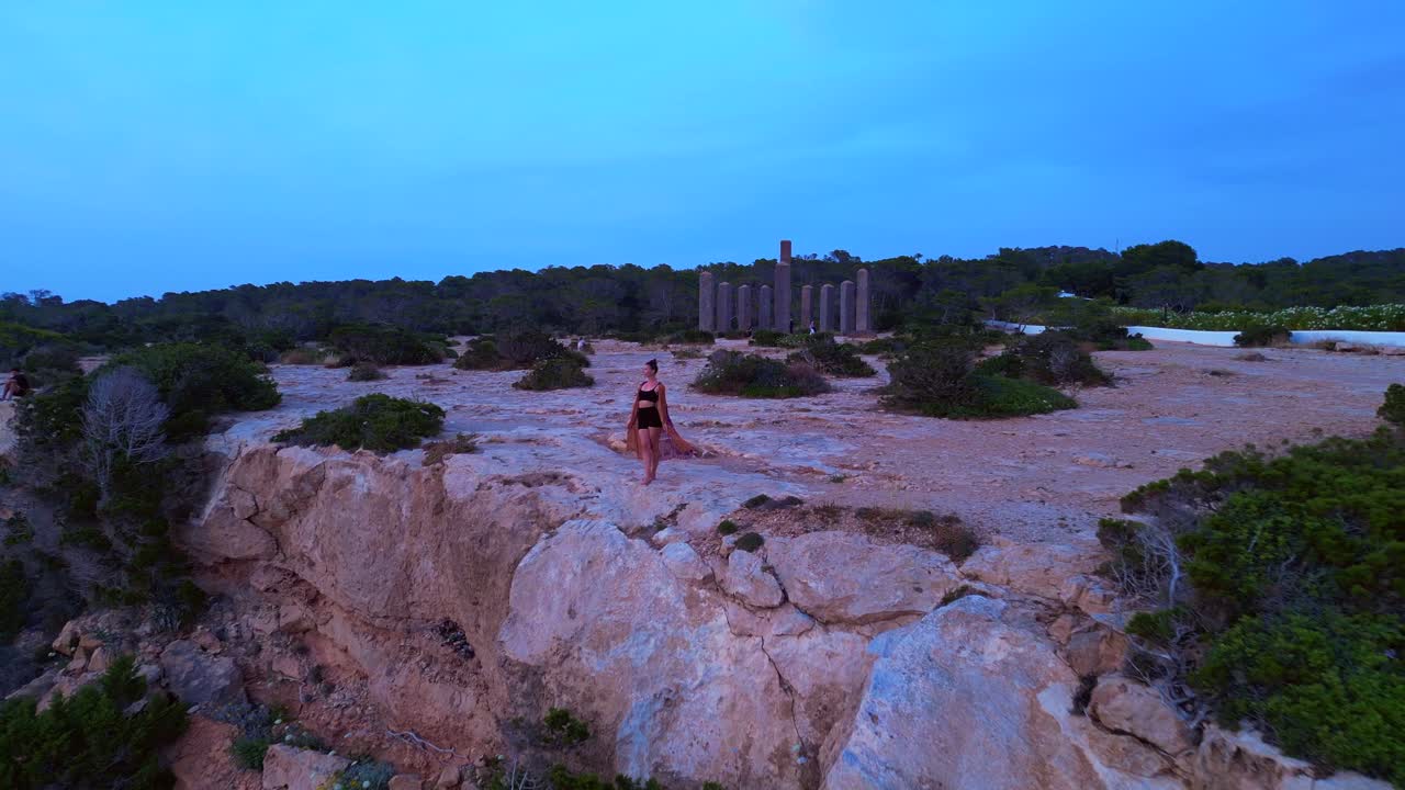 dancing Woman expressing freedom and movement by the sea at a beautiful travel destination Cala Llentia Ibiza. Dramatic aerial view flight fly push forward drone