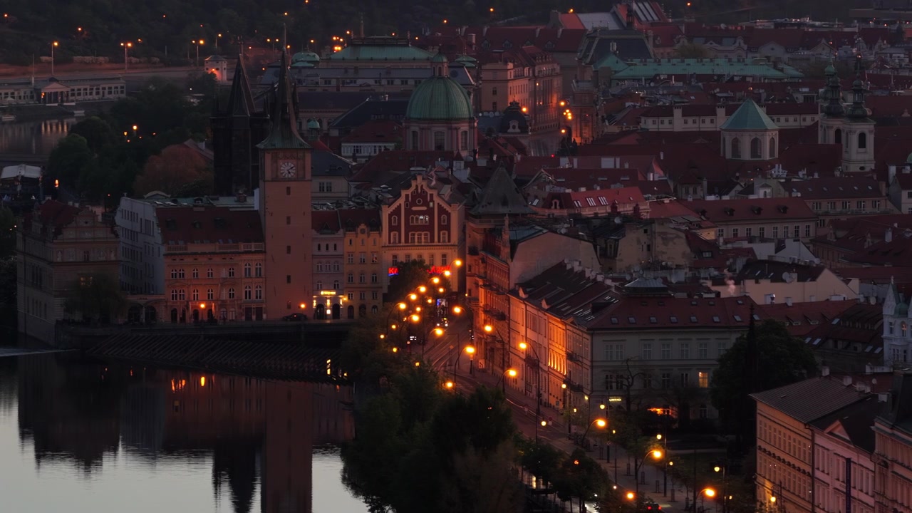 Vista aerea con drone del lungofiume della Città Vecchia con una torre dell'orologio, cupole barocche e tetti rossi riflessi nel fiume Vltava al crepuscolo.