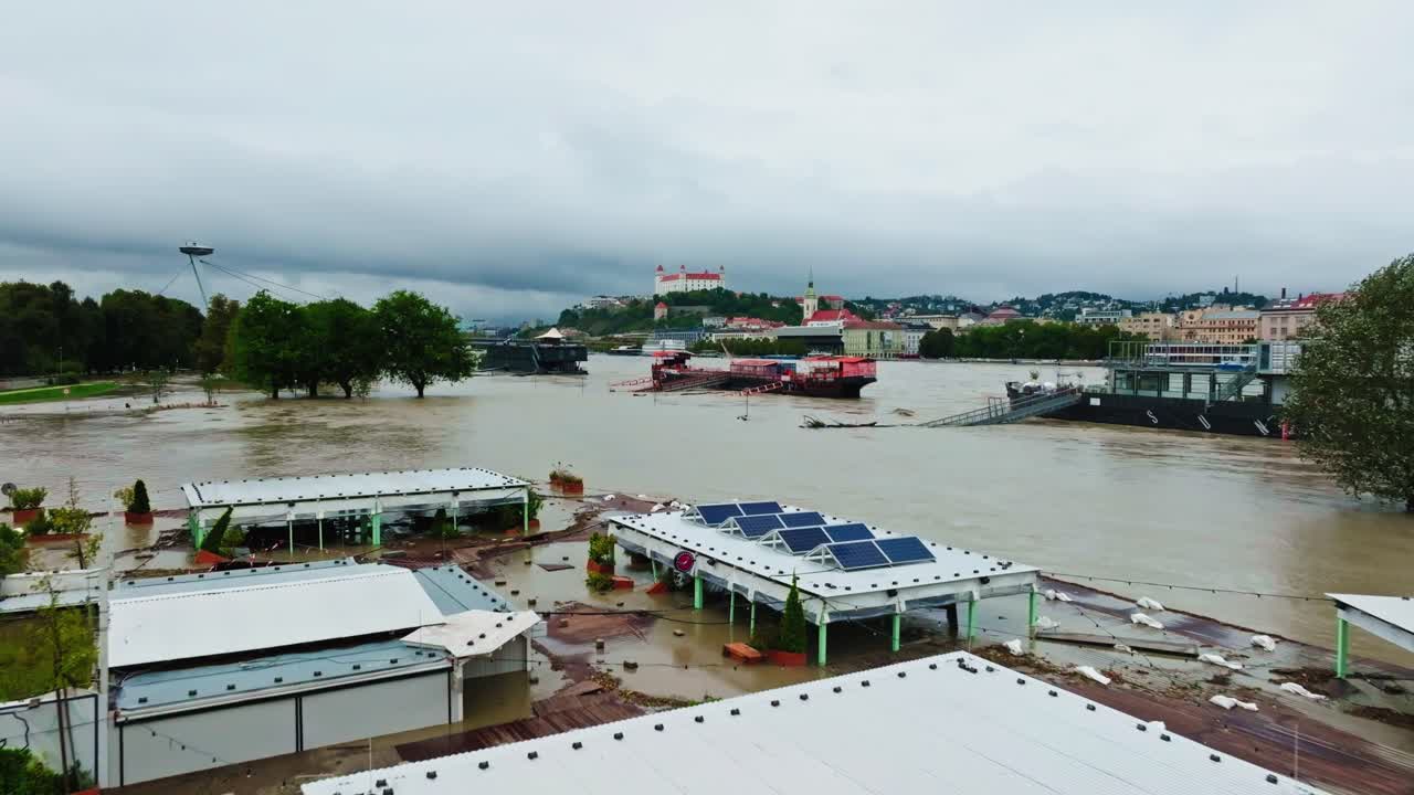 Aerial view of Danube river during a flood. Submerged buildings, walkways, and overflowing water show the extent of the flooding, with the city skyline in the background. (4K)