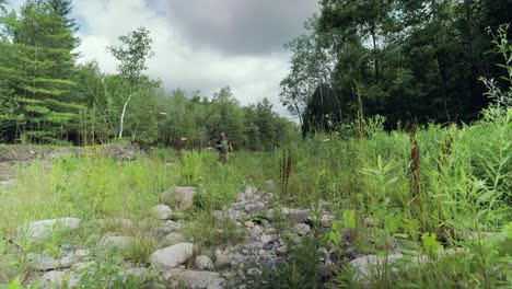 Hiker-with-backpack-wanders-through-rocky-forest-landscape-stepping-over-rocks,-Summer-weather