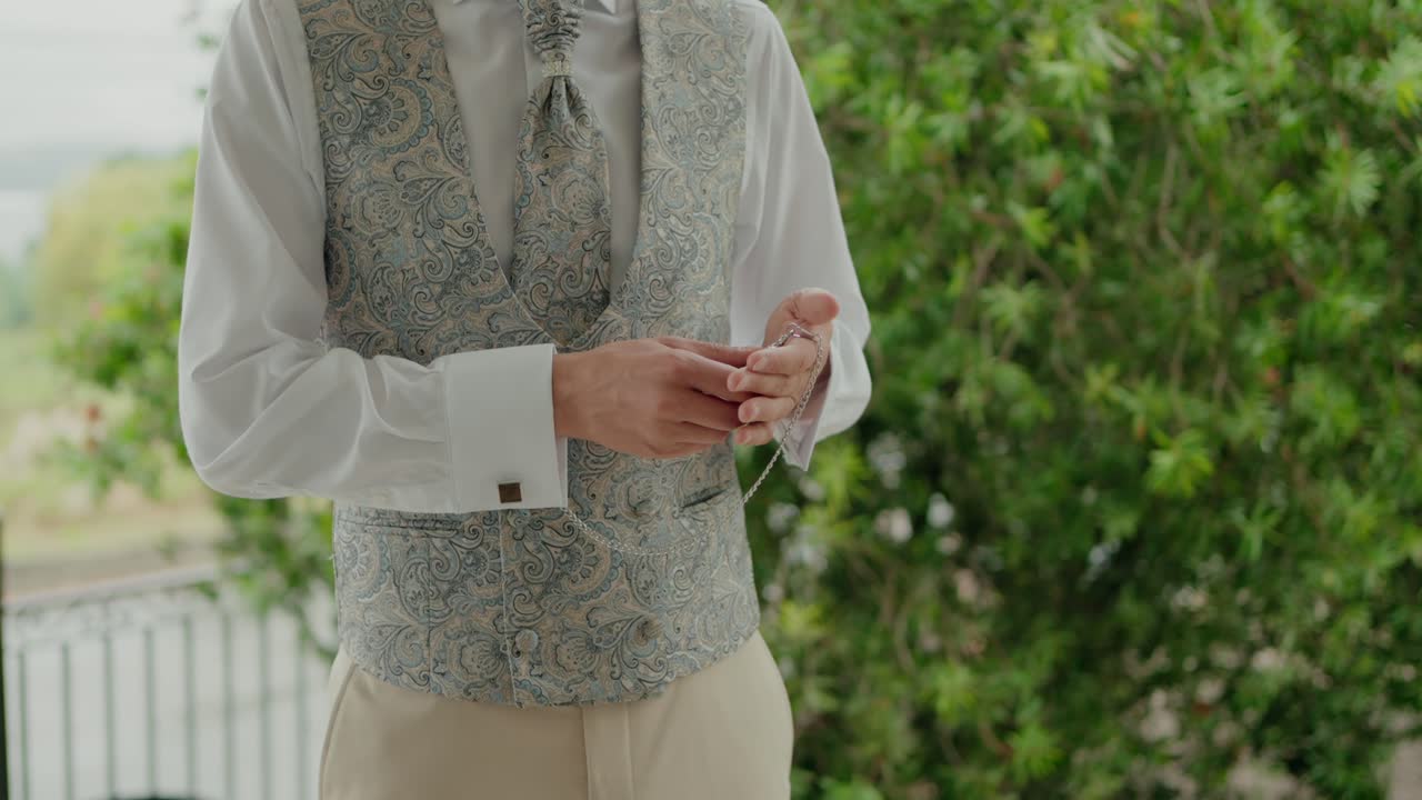 Man holding a pocket watch while wearing a patterned vest, white shirt, and light trousers