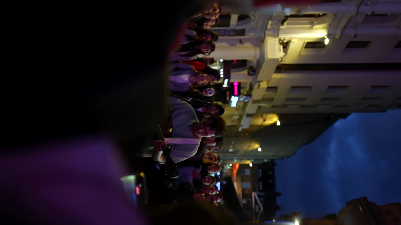 Rack focus shot of busker Myles Crossley performs guitar at night in London's Leicester Square to a crowd of tourists. Vertical
