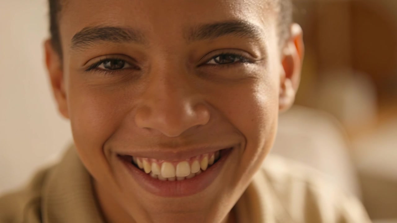 Adolescente sorrindo, abrindo os olhos e ampliando o sorriso enquanto a câmera se mantém próxima na sala de estar, com blusa bege e sofá.