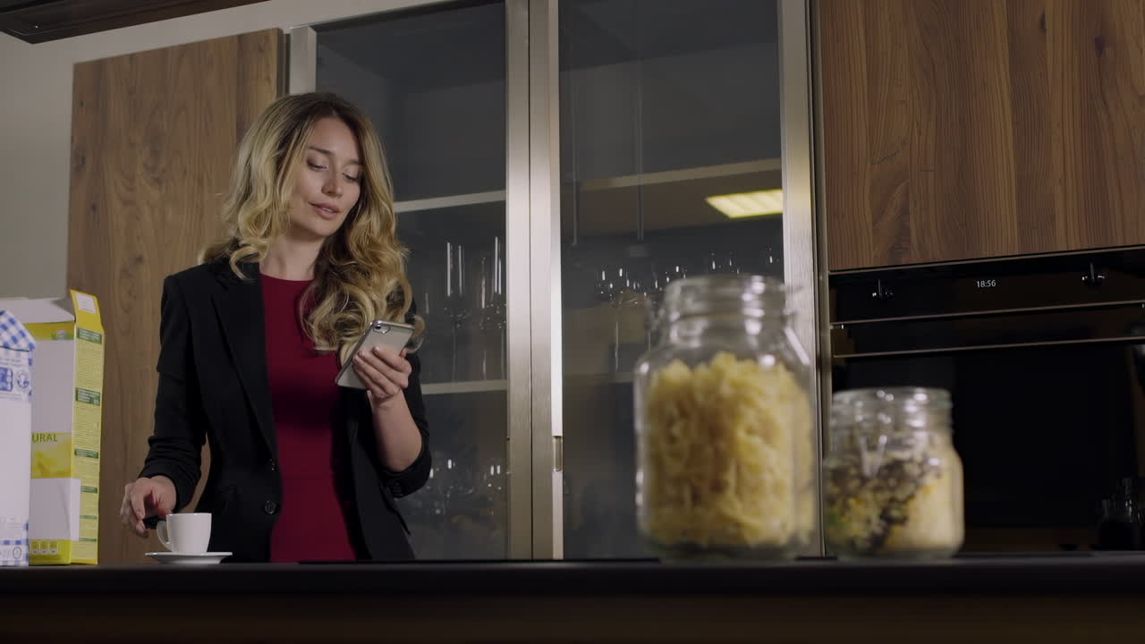 A focused woman stands in a stylish kitchen, using her phone while getting ready to prepare a nutritious snack. Jars of pasta and nuts are placed on the counter