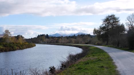 View-of-Caledonian-Canal-and-pathway-with-snow-capped-mountains-in-the-distance-in-Fort-Augustus,-highlands-of-Scotland-UK