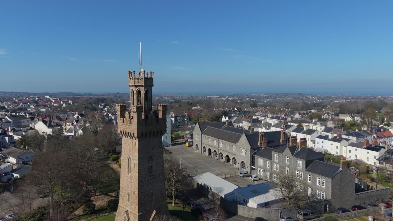 Riprese a bassa quota con drone di St Peter Port Guernsey che volano vicino alla torre dell'Arsenale, mostrando la stazione dei pompieri con viste su tutta l'isola sotto il sole del tardo pomeriggio e un orizzonte nebbioso