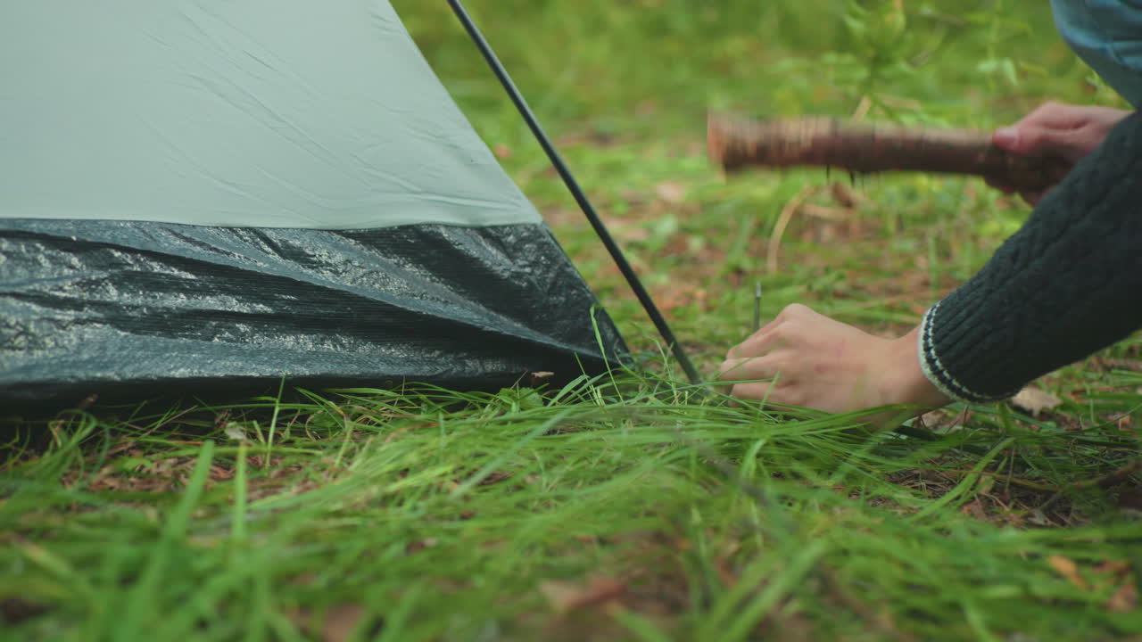 close up showing man hammering tent peg into soft forest ground covered in grass while companion stands nearby holding extra pegs and observing support during secure shelter setup under green canopy