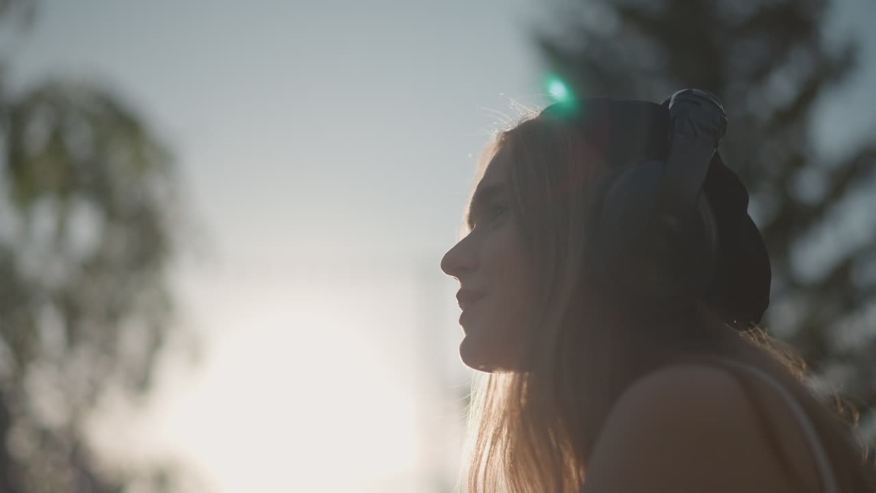 Silueta de mujer con auriculares al atardecer, bebiendo de una botella transparente con luz de fondo cinematográfica y un sutil destello de lente, gorro y auriculares visibles, árboles del parque borrosos al fondo, tonos cálidos de la hora dorada